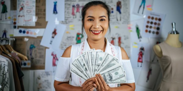 Portrait young asian designer or tailor female standing with a lot of money bank in hand, She smiling with proud in successful of fashion clothes at workshop